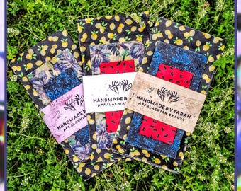 May include: A collection of beeswax and cotton food wraps in various patterns, including lemons, floral designs, and red accents. Each wrap is secured with a branded band that reads "HANDMADE BY TARAN APPALACHIAN BEAUTY". The wraps are displayed on a bed of green foliage.