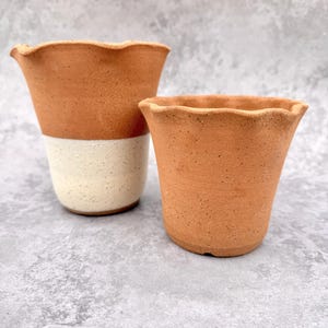 May include: Two ceramic cups with wavy, scalloped rims. The taller cup features a terracotta-coloured upper section and a speckled cream-coloured base. The shorter cup is entirely terracotta-coloured with a speckled texture throughout the surface.