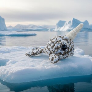 May include: A crocheted narwhal toy with a white horn and black eyes rests on a small iceberg. The toy is primarily white and brown, with a light blue and white background of water and icebergs.