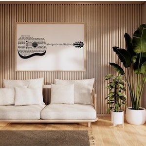 May include: A modern living room with a light-colored sofa, white pillows, and a framed guitar-shaped artwork. Large potted plants with green leaves and a wooden side table add natural elements. The room has wooden paneling and a rug.