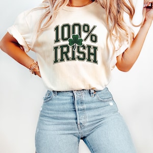 May include: Cream-colored t-shirt with the text "100% Irish" in green block letters and a green shamrock. The shirt is paired with light blue jeans.