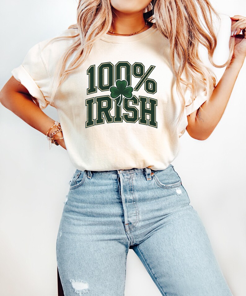 May include: Cream-colored t-shirt with the text "100% Irish" in green block letters and a green shamrock. The shirt is paired with light blue jeans.