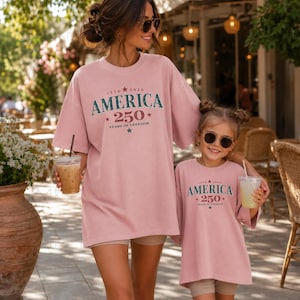 May include: Two matching dusty pink t-shirts featuring dark green and red text that reads AMERICA 250 YEARS OF FREEDOM with stars and the dates 1776 and 2026. The shirts have a crew neckline and short sleeves, designed with a relaxed, loose fit.