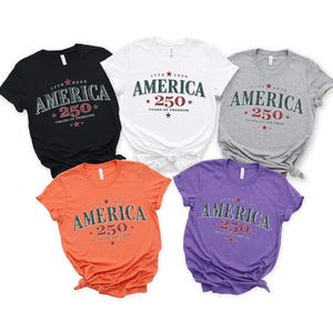 May include: Five short-sleeved t-shirts in black, white, grey, orange, and purple. Each shirt features the text "AMERICA 250 YEARS OF FREEDOM" with "1776-2026" and star graphics printed in a dark green and red color scheme on the chest area.