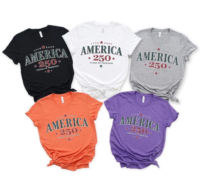 May include: Five short-sleeved t-shirts in black, white, grey, orange, and purple. Each shirt features the text "AMERICA 250 YEARS OF FREEDOM" with "1776-2026" and star graphics printed in a dark green and red color scheme on the chest area.