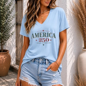 May include: A light blue V-neck t-shirt featuring the text "1776 2026 AMERICA 250 YEARS OF FREEDOM" with red and green stars, paired with light blue denim shorts that have a distressed, frayed hemline and visible rips on the front panels.