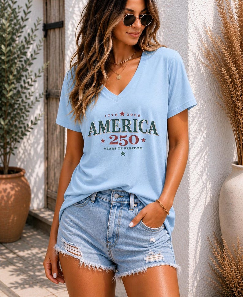 May include: A light blue V-neck t-shirt featuring the text "1776 2026 AMERICA 250 YEARS OF FREEDOM" with red and green stars, paired with light blue denim shorts that have a distressed, frayed hemline and visible rips on the front panels.