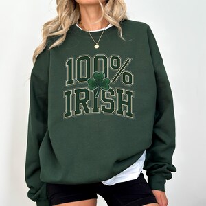 May include: Dark green sweatshirt with the text "100% Irish" in a white and gold font, featuring a green shamrock. The sweatshirt has long sleeves and a crew neck. The model is wearing black shorts and a white undershirt.