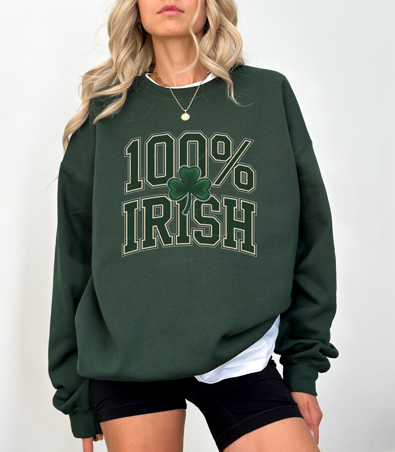 May include: Dark green sweatshirt with the text "100% Irish" in a white and gold font, featuring a green shamrock. The sweatshirt has long sleeves and a crew neck. The model is wearing black shorts and a white undershirt.