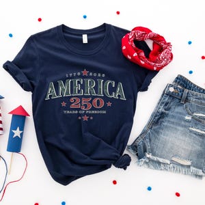 May include: A navy blue t-shirt featuring the text AMERICA 250 YEARS OF FREEDOM with red and white stars, paired with blue denim shorts and a red patterned bandana. Two paper rocket decorations with red and white stripes are also present.