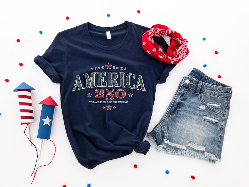May include: A navy blue t-shirt featuring the text AMERICA 250 YEARS OF FREEDOM with red and white stars, paired with blue denim shorts and a red patterned bandana. Two paper rocket decorations with red and white stripes are also present.