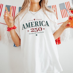 May include: A white short-sleeve t-shirt featuring the dark green and red text AMERICA 250 YEARS OF FREEDOM with the years 1776 and 2026 and small star graphics printed on the front center of the garment in a collegiate style font.