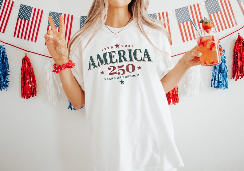 May include: A white short-sleeve t-shirt featuring the dark green and red text AMERICA 250 YEARS OF FREEDOM with the years 1776 and 2026 and small star graphics printed on the front center of the garment in a collegiate style font.