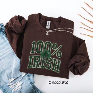 May include: A chocolate-colored sweatshirt with the text "100% Irish" in green and white, featuring a shamrock. A silver necklace is draped across the shirt. The word "Chocolate" is written below the shirt. The sweatshirt is paired with blue jeans.
