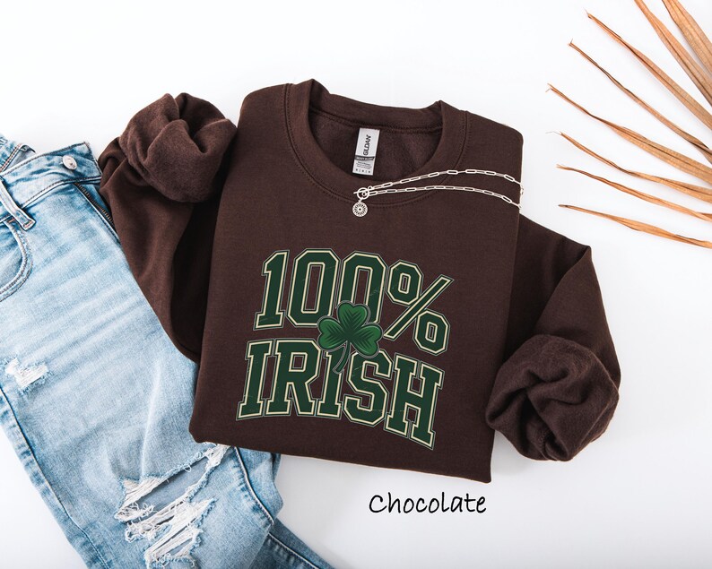 May include: A chocolate-colored sweatshirt with the text "100% Irish" in green and white, featuring a shamrock. A silver necklace is draped across the shirt. The word "Chocolate" is written below the shirt. The sweatshirt is paired with blue jeans.