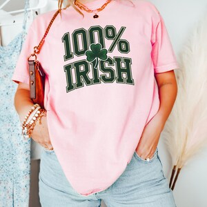 May include: A light pink t-shirt with the text "100% Irish" in dark green, featuring a shamrock. The shirt is paired with light blue jeans. A gold necklace and bracelets are visible.