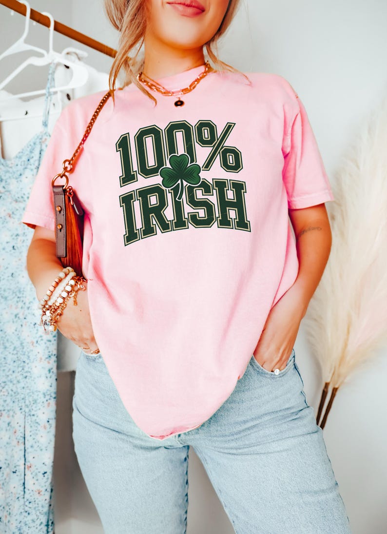 May include: A light pink t-shirt with the text "100% Irish" in dark green, featuring a shamrock. The shirt is paired with light blue jeans. A gold necklace and bracelets are visible.