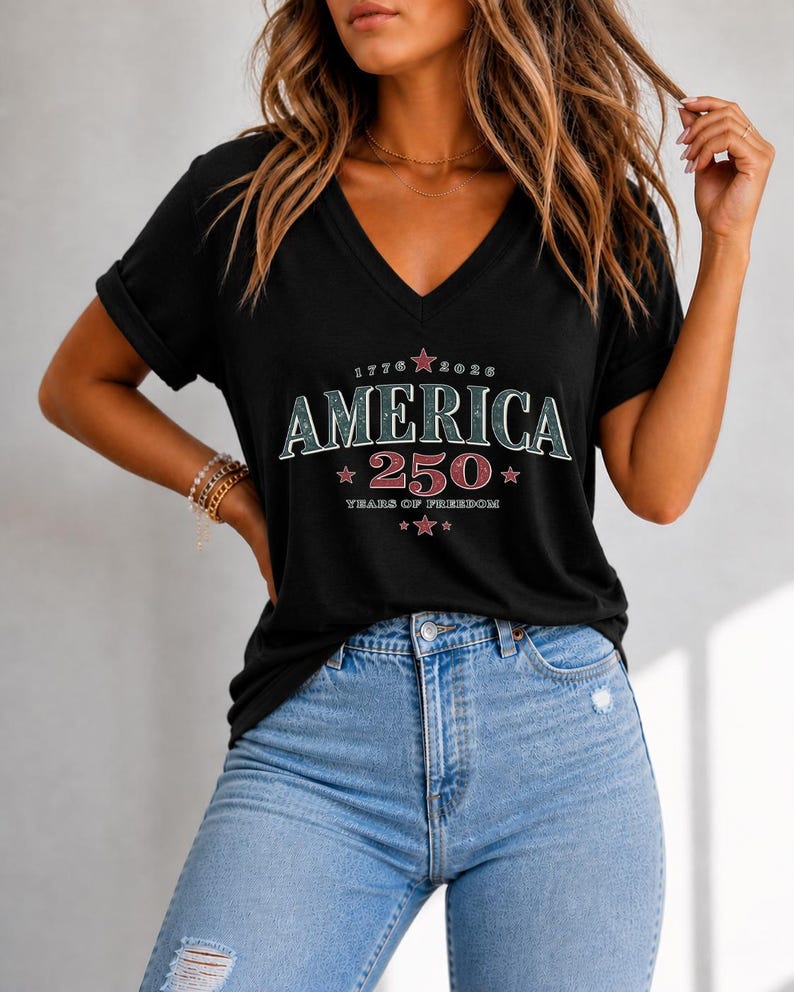 May include: A black V-neck t-shirt with rolled sleeves featuring a graphic print that reads AMERICA 250 YEARS OF FREEDOM 1776 2026 with red stars. The shirt is paired with light blue denim jeans that have a distressed detail on the upper thigh area.