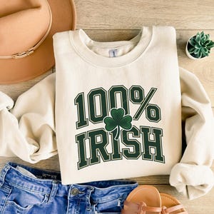 May include: Cream-colored sweatshirt with the text "100% Irish" in green, featuring a shamrock. The sweatshirt is paired with blue denim shorts, a brown hat, and sandals. A small succulent plant in a white pot is also visible.