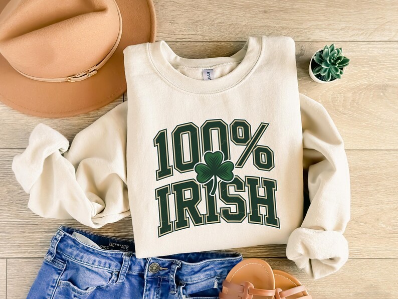 May include: Cream-colored sweatshirt with the text "100% Irish" in green, featuring a shamrock. The sweatshirt is paired with blue denim shorts, a brown hat, and sandals. A small succulent plant in a white pot is also visible.