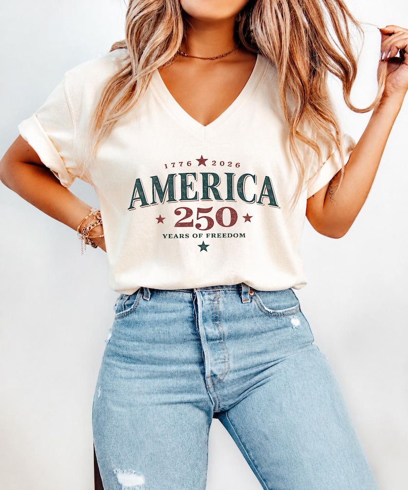 May include: A cream-colored V-neck t-shirt featuring the text 1776 2026 AMERICA 250 YEARS OF FREEDOM in dark green and maroon, accented with stars. The shirt is paired with light blue denim jeans that have distressed detailing on the thighs.