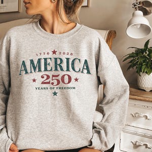 May include: A light grey crewneck sweatshirt featuring a graphic print with the text 1776 2026 AMERICA 250 YEARS OF FREEDOM. The design includes red and teal stars and lettering with a distressed texture across the center of the chest.