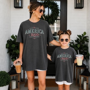 May include: Two charcoal gray t-shirts featuring a vintage-style graphic with the text "AMERICA 250 YEARS OF FREEDOM" in red and white lettering, accented with small red stars. The shirts have a relaxed fit and short sleeves, worn by two individuals.