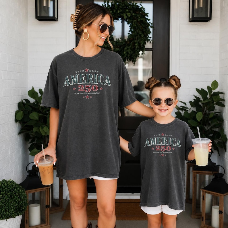 May include: Two charcoal gray t-shirts featuring a vintage-style graphic with the text "AMERICA 250 YEARS OF FREEDOM" in red and white lettering, accented with small red stars. The shirts have a relaxed fit and short sleeves, worn by two individuals.