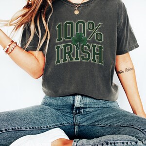 May include: Dark gray t-shirt with the text "100% IRISH" in green and white, featuring a green shamrock. The shirt is a casual style, suitable for St. Patrick's Day or expressing Irish heritage.