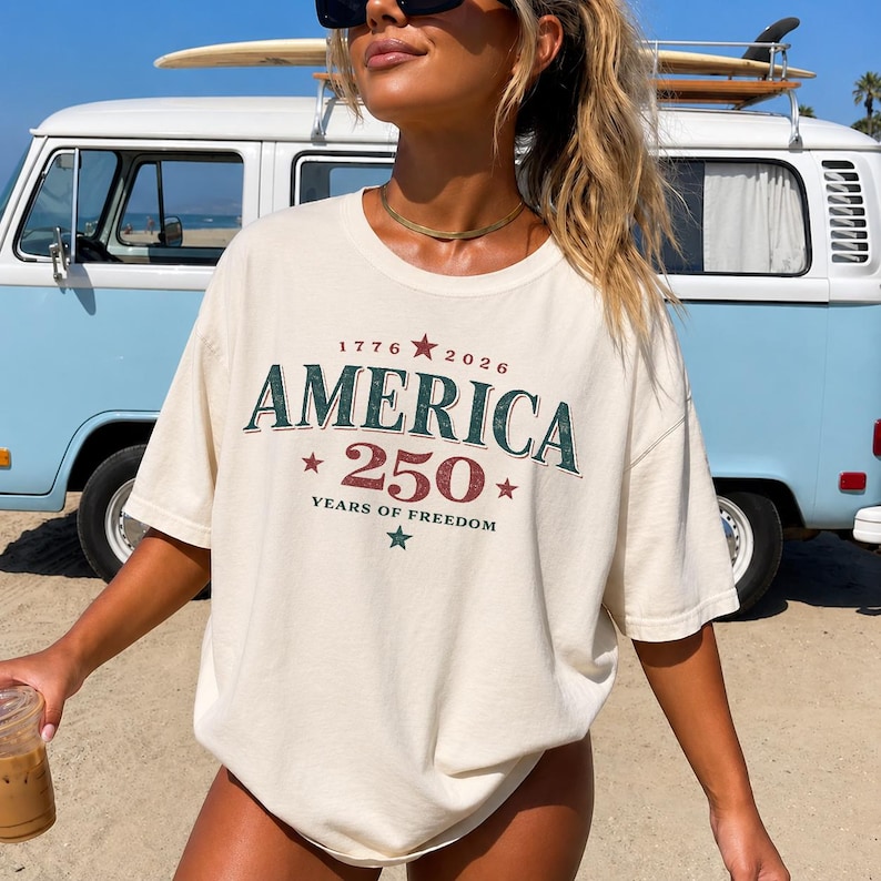 May include: A cream-colored short-sleeve t-shirt featuring a distressed graphic print. The text reads 1776 2026 AMERICA 250 YEARS OF FREEDOM, accented with red and green stars. The design has a vintage, weathered appearance with bold lettering.