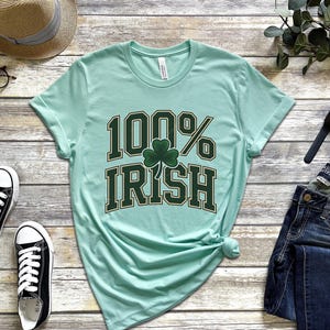May include: A light green t-shirt with the text "100% Irish" in dark green lettering and a shamrock graphic. The shirt is styled with blue jeans, black sneakers, a straw hat, and eyeglasses.