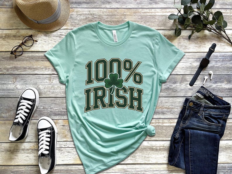 May include: A light green t-shirt with the text "100% Irish" in dark green lettering and a shamrock graphic. The shirt is styled with blue jeans, black sneakers, a straw hat, and eyeglasses.