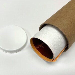 May include: A brown cardboard tube with a white lid. The tube is partially unrolled, revealing a rolled-up poster with orange, red, and green colors.