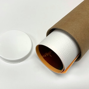 Puede incluir: Una impresi&oacute;n art&iacute;stica enrollada con una superficie blanca texturizada es parcialmente visible dentro de un tubo de cart&oacute;n marr&oacute;n. Una tapa de pl&aacute;stico blanco se encuentra junto al tubo. La impresi&oacute;n tiene acentos naranjas y verdes.