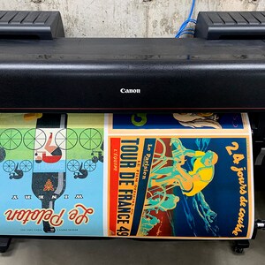 May include: A black Canon imagePROGRAF large format printer with a roll of paper feeding through it. The paper has three vintage-style posters printed on it, featuring cyclists and the text "Tour de France 49" and "24 jours de course".
