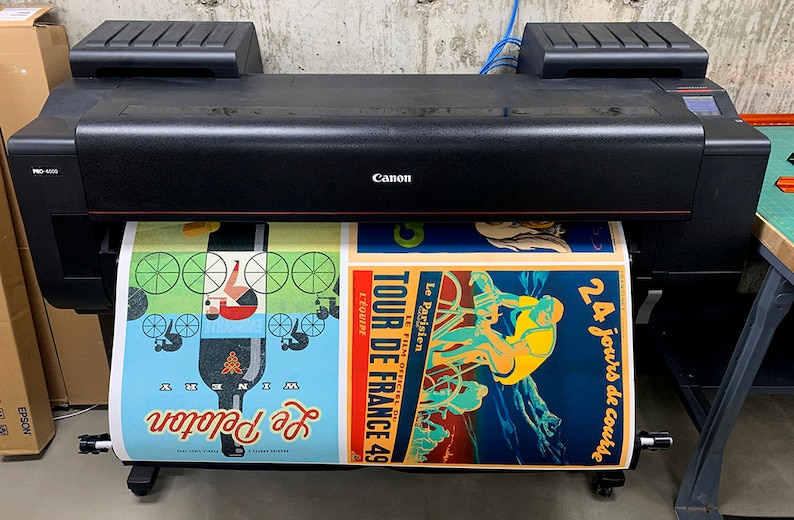May include: A black Canon imagePROGRAF large format printer with a roll of paper feeding through it. The paper has three vintage-style posters printed on it, featuring cyclists and the text "Tour de France 49" and "24 jours de course".
