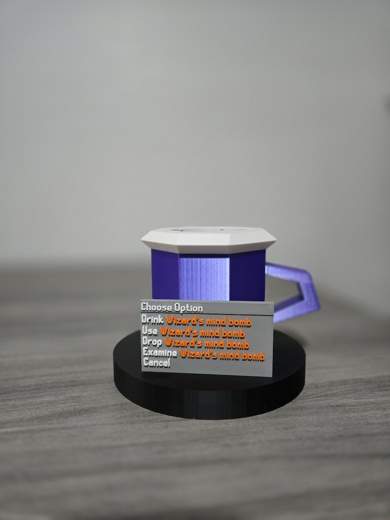 May include: A 3D-printed mug with a purple body, white top, and a handle. The mug sits on a black circular base. A small rectangular sign displays the text "Choose Option" and other options related to "Wizard's mind bomb."