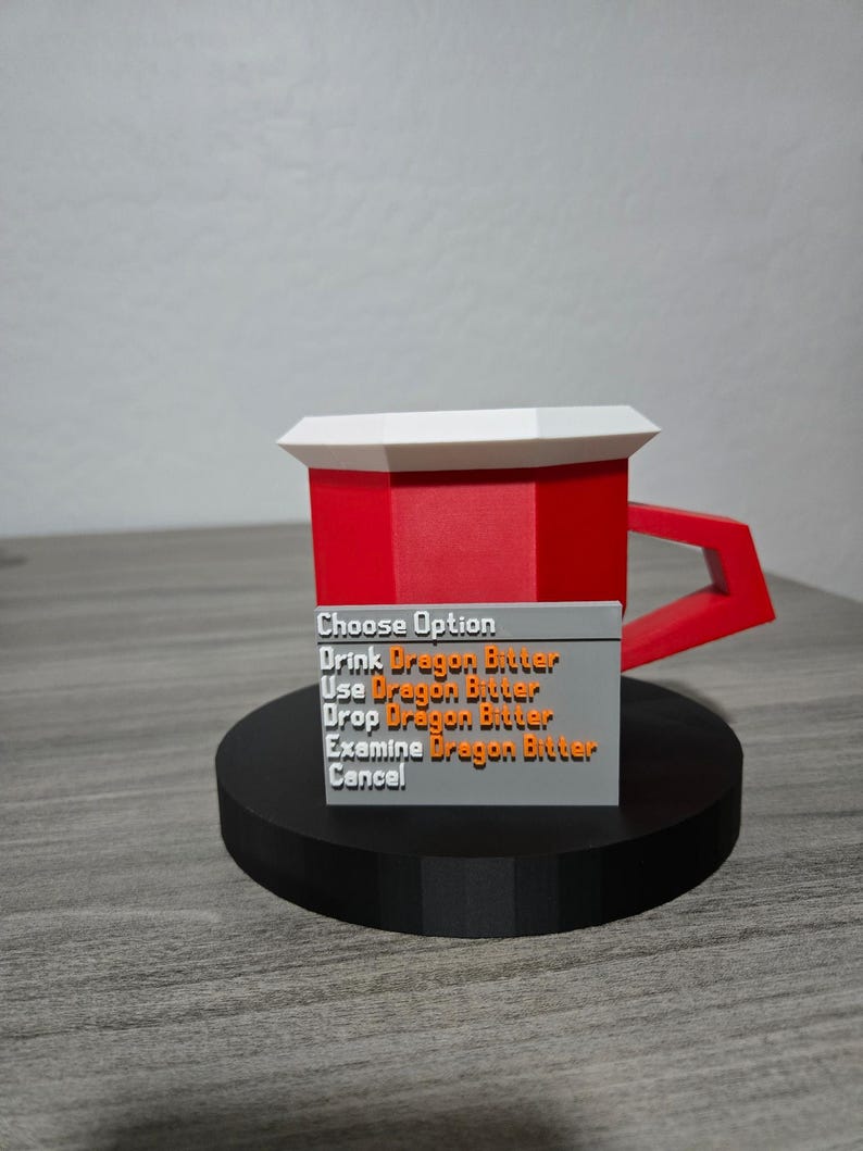 May include: A red and white mug with a geometric design, featuring a handle and a base. A grey rectangular sign displays the text "Choose Option" and several options related to "Dragon Bitter". The mug is on a black circular base.