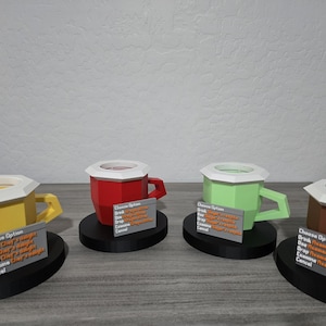 May include: Four brightly coloured, geometric mugs with white rims and handles, each on a black base. The mugs are yellow, red, green, and brown. Each mug has a small sign with text. The mugs are displayed on a grey surface.