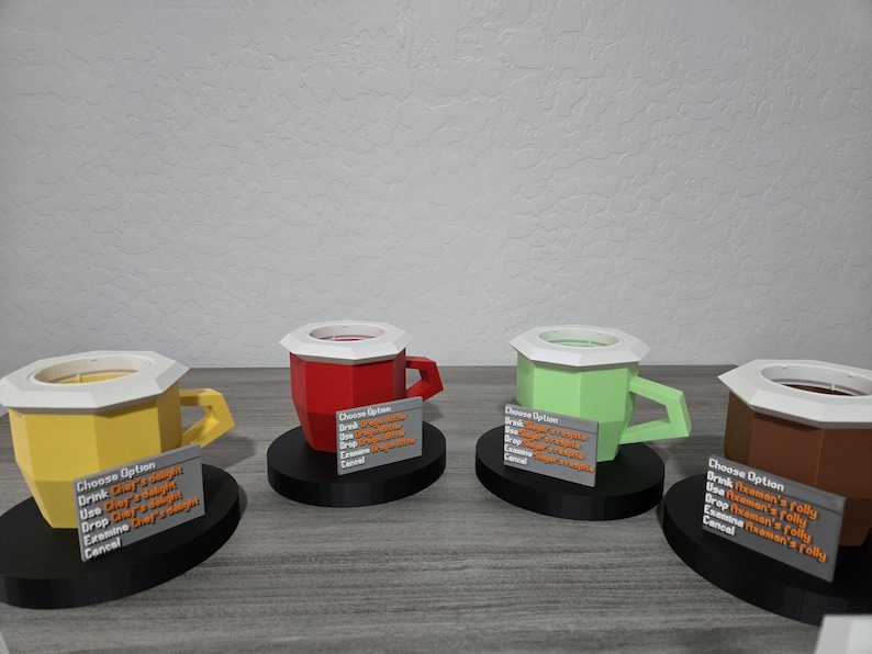May include: Four brightly coloured, geometric mugs with white rims and handles, each on a black base. The mugs are yellow, red, green, and brown. Each mug has a small sign with text. The mugs are displayed on a grey surface.