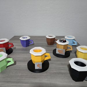 May include: A selection of colourful, geometric paper coffee cups with white rims and handles. The cups are in various colours, including red, green, yellow, blue, brown, and black. Some cups have a black base.
