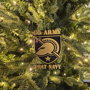 Puede incluir: Adorno navideño con las palabras doradas "GO ARMY BEAT NAVY". El adorno tiene forma de escudo con un diseño dorado de casco y espada sobre fondo negro. Cuelga de un árbol de Navidad verde con luces blancas.