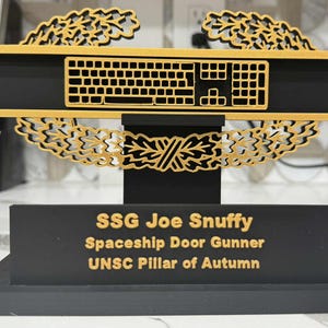 Pode incluir: Um prêmio decorativo preto e dourado com um design de teclado. O prêmio tem uma base retangular com o texto "SSG Joe Snuffy Spaceship Door Gunner UNSC Pillar of Autumn". O teclado é dourado, e o texto é dourado.