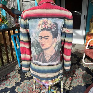 May include: A colourful blazer with a Frida Kahlo portrait on the back. The jacket has a striped pattern in red, blue, and green. The portrait is framed by a black border.