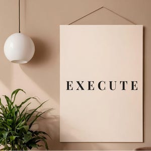 Puede incluir: Una impresión artística de pared minimalista con la palabra "EXECUTE" en negrita, fuente serif negra. La impresión es de color crema y cuelga de una pared de tonos neutros. También se ve una lámpara colgante esférica blanca y una planta verde.