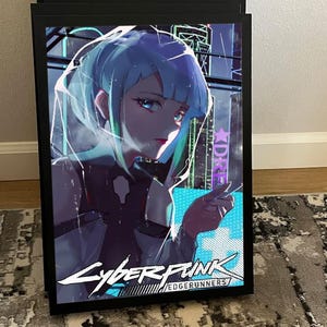 K&ouml;nnte beinhalten: Ein gerahmtes Poster mit einer Cyberpunk-Anime-Illustration. Das Kunstwerk zeigt eine weibliche Figur mit blau-gr&uuml;nen Haaren vor einer futuristischen Stadtlandschaft. Das Poster enth&auml;lt den Text "Cyberpunk Edgerunners" in einer stilisierten Schriftart, in einem schwarzen Rahmen.