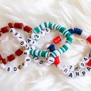 May include: Three beaded bracelets in red, white, blue, and green. One bracelet has the letters "RANP" and numbers. The other bracelets have numbers. The bracelets are made of small, colorful beads and white letter beads.