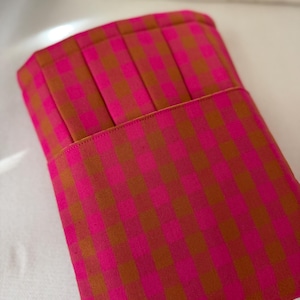 May include: A rectangular fabric sleeve featuring a vibrant hot pink and olive green gingham check pattern. The top section of the sleeve is constructed with vertical pleats, while the lower portion remains flat with a clean stitched edge.