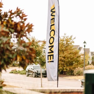 May include: A white flag with the word "WELCOME!" in yellow lettering. The flag is displayed outdoors, with a blurred background of trees, a street, and a car. The flag is attached to a pole.