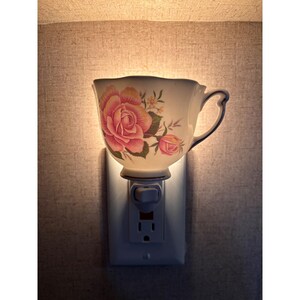 May include: A wall-mounted plug-in night light featuring a white porcelain teacup with a scalloped rim and a handle. The teacup is decorated with a large pink rose and a smaller rosebud with green leaves. The light emits a warm glow from within.